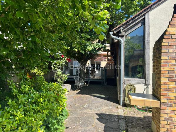 À proximité immédiate du centre-ville, des gares, des axes autoroutiers et de la tangentielle, cette authentique maison des années 1930 séduira les amoureux de cachet, d’espaces chaleureux et de projets de vie polyvalents.

Dès l’entrée, un couloir distribue : • une première chambre, • un toilette indépendant avec lave-mains, • une seconde chambre.
La visite se poursuit vers une cuisine récente, aménagée et équipée, ouverte sur une superbe extension baignée de lumière. Cette pièce de vie au charme incomparable offre une cheminée, une belle hauteur sous plafond et de magnifiques poutres apparentes. Grâce à ses larges ouvertures sur le jardin, l’intérieur et l’extérieur se répondent harmonieusement tout au long de l’année.

À l’étage, un palier aménagé en coin bibliothèque dessert : • une chambre, • une salle d’eau avec toilette, lavabo et bidet.
La maison bénéficie également d’un sous-sol complet comprenant plusieurs caves, une chaufferie ainsi qu’une pièce de 25 m² pouvant être aménagée en espace habitable, bureau indépendant, salle de loisirs ou studio selon vos besoins.

À l’extérieur, le jardin intimiste et arboré accueille : • un atelier indépendant d’environ 40 m², • deux garages de plus de 16 m² chacun, situés au fond du terrain, • une agréable terrasse ombragée par un magnifique tilleul.
Ces dépendances offrent un véritable potentiel pour une activité professionnelle ou un investissement locatif : atelier d’artiste, cabinet de profession libérale, espace de coworking, activité artisanale, stockage, studio indépendant ou bureaux.
Cette propriété conviendra parfaitement : • à un artisan souhaitant disposer d’un atelier à domicile, • à un artiste ou créatif recherchant un espace de travail indépendant, • à un professionnel libéral (architecte, thérapeute, coach, consultant, profession paramédicale…), • ou encore à un investisseur désirant exploiter les dépendances pour du locatif ou une activité complémentaire.

Une maison pleine d’âme et de possibilités, idéale pour conjuguer vie personnelle, activité professionnelle et projet patrimonial.

Taxe foncière: 2268€
Energie: 238€/mois