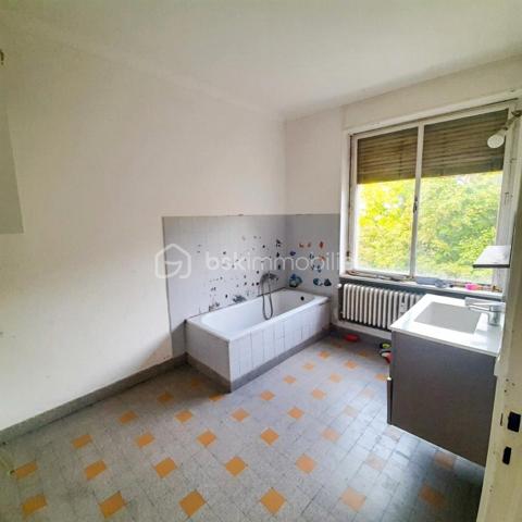 Situé au 3ᵉ étage du bâtiment annulaire, rue Auguste-Wicky à Mulhouse, cet appartement de 92 m² bénéficie d'un emplacement privilégié, à proximité immédiate de la gare et à deux pas du centre historique. Ses beaux volumes et sa configuration offrent de nombreuses possibilités d'aménagement.

Une adresse emblématique de Mulhouse
Construit dans les années 1950 par l'architecte Pierre-Jean Guth, le bâtiment annulaire est un édifice singulier de l'architecture mulhousienne d'après-guerre. Sa forme circulaire remarquable et son histoire lui ont valu une inscription au titre des monuments historiques en 2006. Une adresse de caractère, au cœur d'un ensemble architectural qui fait partie du patrimoine de la ville.

Un appartement à aménager selon votre projet
L'appartement se compose d'une large entrée, de quatre belles pièces de 11, 12, 16 et 17 m², offrant différentes possibilités d'aménagement selon les besoins de chacun. Une cuisine indépendante, une salle de bains, un WC séparé et un petit balcon complètent le bien.
Des travaux de rénovation sont à prévoir, laissant la possibilité de repenser les espaces et les prestations selon vos envies. Un devis a déjà été réalisé afin de permettre aux acquéreurs d'anticiper le budget nécessaire à leur projet.
À l'arrière de l'immeuble, la cour intérieure verdoyante entretenue par la Ville de Mulhouse apportent une touche de nature en milieu urbain. Le bien dispose également d'une cave. Des possibilités de stationnement existent à l'arrière de l'immeuble, sous réserve de disponibilité.

Que ce soit pour créer un lieu de vie personnalisé ou pour réaliser un investissement locatif, cet appartement présente un potentiel intéressant. Le règlement de copropriété autorise la location de courte durée, un atout supplémentaire pour les acquéreurs souhaitant étudier cette possibilité, sous réserve du respect de la réglementation applicable.

Informations complémentaires
- Surface : 92 m²
- Étage : 3ᵉ
- Chauffage collectif au gaz
- DPE : D — GES : D
- Taxe foncière : 1 200 € par an
- Charges annuelles comprennant charges générales de l'immeuble, eau chaude, eau froide et chauffage (5830€ de charges locatives sur 6283€ en 2025)