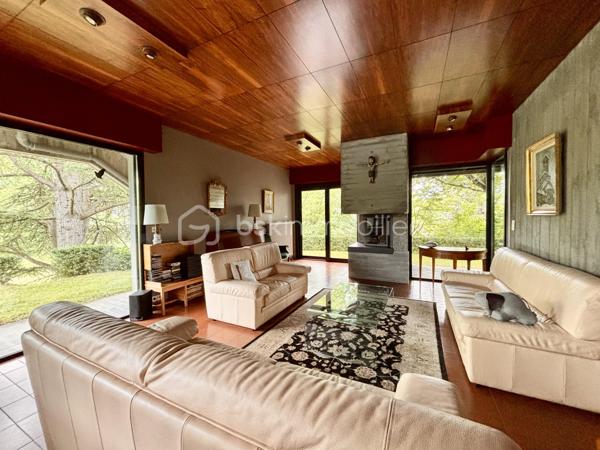 bonjour. Voici l'essentiel : 
à Gières au fort du Mûrier, une villa-belvédère 
258 m2 habitables 
158 m2 locaux annexes
416 m2 surface utile
6 chambres
4 salles de bain
Piscine à débordement
Terrain arboré et clos : 6737 m2 (chemin de ronde partiellement)

Bienvenue à celles et ceux qui préfère emprunter le « chemin de ronde » architectural :
A l'aube des années 80, aucune entrave à l'audace de ces jeunes architectes et commanditaires qui saisissent cette exceptionnelle opportunité de construire cette villa-belvédère au flanc du Fort du Mûrier à Gières qui verrouille la vallée du Grésivaudan.
La villa lui emprunte sa matérialité, son ancrage et sa compacité qu'elle exprime par les outils architecturaux développés dans le sillage de Le Corbusier.
Là où le Fort retourne comme un gant l'alignement des façades de ses casemates en une rue utilitaire  encastrée, la villa, sous le ruban de la haute poutre-acrotère du toit-terrasse, déploie, par redents irréguliers, l'alternance de ses parois pleines et vitrées toute hauteur.
Elle exalte la minéralité de sa périphérie de terrasses gravillonnées, d'une brique nuancée appareillée horizontalement en pans ourlés et d'un béton aux épidermes multiples tantôt lisse, tantôt brut de décoffrage, parfois avec méplats mais toujours d'expression verticale et favorise sa vocation résidentielle par les nombreux et généreux pans vitrés de son rez-de-jardin de 222 m2 intégralement de plain-pied. 
A cette projection dans le paysage proche et lointain répond une fluidité intérieure autorisée par la prédilection des échappées visuelles obliques et un certain dépouillement général.
Au final,  sans en imposer mais avec fermeté, sans mimétisme au fort, par son inaltérable minimalisme, la villa-belvédère semble surgie hier autant que depuis des temps immémoriaux.

Une chambre d'écho aux bruissements des éléments, du végétal et de la montagne.
Comme le fort organise sa vie défensive en fonction de sa rue en boomerang, la villa répartit son organisation de part et d'autre d'une rue intérieure dotée de la même orientation nord-ouest/sud-est salutaire eu égard au réchauffement climatique. 
Au nord, l’espace nuit de hauteur conventionnelle comprend 4 chambres, 3 salles d’eau nimbées d'une subtile lumière zénithale et 2 wc. Toutes les chambres s'exonèrent d'une vue frontale sur le fort au profit d'orientations est et ouest plus agréables. La suite parentale outre le privilège de ses 27 m2 et sa baie cadrant les trois pics de Freydone, dispose d'un accès doublé par le salon surélevé. Arrêtons-nous sur cette pièce singulière qui jouit d'un rapport visuel immédiat avec le miroir d'eau sur lequel flotte la chaine de Belledonne...salon tour-à-tour empli de la rumeur ludique de la piscine et assurément contemplative la nuit ou aux aurores, sa triple ouverture et son surplomb du hall l'apparentent au campement à la « croisée des chemins »  ce dont atteste la ligne fuyant et sinueuse des ganivelles de châtaigner du chemin de ronde.

Au sud de la rue intérieure, avec laquelle ils partagent une hauteur sous plafond avoisinant les 3 mètres, se déploient les lieux de rassemblement ou de concentration que sont le séjour, la cuisine, la salle-à-manger et le bureau.
Le séjour en contrebas de la rue intérieure s'organise autour du bloc massif intégrant le foyer-insert et son alter-ego le barbecue de la terrasse couverte des repas de mi-saison et d'été. Son plafond de panneaux de chêne massif confère à la pièce une couleur acoustique propice aux confidences.
On peut qualifier la cuisine d'agora familiale tant son organisation centripète permet de s'y tenir bien au-delà de sa fonction première de préparation des repas. Sa table tout en transparence dissout la frontière entre cuisine et terrasse tandis que ses ouvertures cardinales en diagonale permettent un contrôle visuel de toute la propriété : l'arrivée des visiteurs, le chevreuil du chemin de ronde, le gazouillis des oiseaux proches. Et que dire de son mobilier minimaliste et inaltérable aux couleurs hédoniques qui répond à sa mission depuis un demi-siècle avec la même justesse ?
Le niveau bas -d'égale surface- est purement fonctionnel. Au nord-ouest et de plain-pied avec l'accès à la propriété, le vaste garage est accessible par 3 portes sectionnelles et donne accès à un studio. Du fait de l'encastrement dans la déclivité du terrain et donc totalement enterrés, vous trouvez les espaces techniques tels que -principalement- la filtration de la piscine à débordement, la chaufferie et sa chaudière Weishaupt, la conserverie-cave à vins, la buanderie, l'atelier. Les vides sanitaires hauteur d'homme et partiellement soustrait à la roche réservent des opportunités d'aménagement. 

La villa s'implante au sud de l'équerre formé par le chemin de ronde et en lisière du hameau -solitaire mais solidaire- dont elle se protège par un écran végétal et une certaine déclivité.
Prolongement naturel du demi-kilomètre que constitue le tour de la propriété, un escalier extérieur donne accès au toit-terrasse qui offre une vue panoramique à 360 degrés sur le Vercors, Grenoble, la dent de Crolles, le Grésivaudan, le Mont-blanc par beau temps et la chaine de Belledonne. 
La boucle du Fort du Mûrier passe au pied de la propriété et offre au cours de ses deux heures de marche une succession de panoramas, selon les randonneurs, « à coupe le souffle ».
Aux portes de Grenoble, Gières, bien desservie par la rocade nord-est et à la croisée de plusieurs domaines skiables rejoints en moins d'une demie-heure, elle est dotée de tous les commerces et services. Mais une telle adresse se mérite et il faut - depuis le bourg - gravir un dénivelé de plus de 200 mètres pour atteindre le Fort et la villa.

Alain CHIAUDANO & Régis HERBIN, l’héroïque binôme architectural 
Durant les deux années précédant la construction, les commanditaires ont affronté de nombreuses résistances à leur projet mais cela n'a entamé en rien ni leur envie de vivre en ce lieu solitaire et grandiose ni la conviction d'Alain Chiaudano d'un plateau de vie -« bunker » en lévitation- affranchi de toute enveloppe conventionnelle. Malheureusement, quelques mois avant l'ouverture du chantier, l'architecte est emporté par la maladie et c'est son ami et associé régis Herbin qui fera preuve d'abnégation et de respect mémoriel. Porté par l'encouragement sans faille des commanditaires, le jeune architecte livrera en 15 mois une œuvre unique où tout est calepiné, apparent, doté d'une science des rythmes, des épidermes et de l'espace qui n'est pas sans rappeler l'Atelier de Montrouge de la première heure ou le binôme Le Corbusier-Wogenscky. 
Les améliorations que les résidents lui ont prodigué au fil des ans, la belle patine des ouvrages et au final, l'absence de toute modification majeure témoignent de la pertinence des choix initiaux et attestent de son état impeccable.