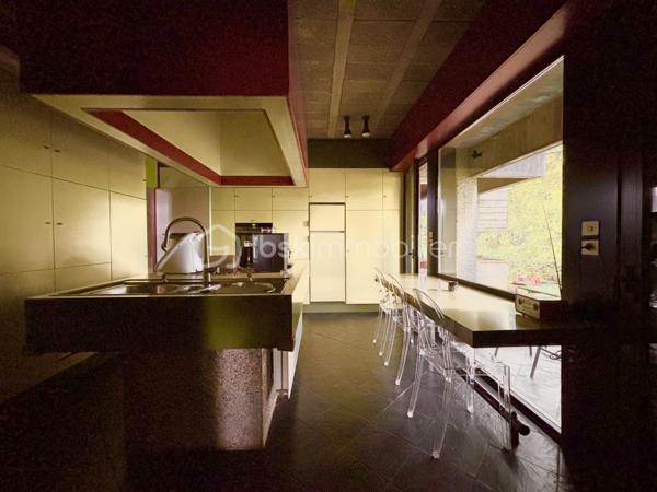 bonjour. Voici l'essentiel : 
à Gières au fort du Mûrier, une villa-belvédère 
258 m2 habitables 
158 m2 locaux annexes
416 m2 surface utile
6 chambres
4 salles de bain
Piscine à débordement
Terrain arboré et clos : 6737 m2 (chemin de ronde partiellement)

Bienvenue à celles et ceux qui préfère emprunter le « chemin de ronde » architectural :
A l'aube des années 80, aucune entrave à l'audace de ces jeunes architectes et commanditaires qui saisissent cette exceptionnelle opportunité de construire cette villa-belvédère au flanc du Fort du Mûrier à Gières qui verrouille la vallée du Grésivaudan.
La villa lui emprunte sa matérialité, son ancrage et sa compacité qu'elle exprime par les outils architecturaux développés dans le sillage de Le Corbusier.
Là où le Fort retourne comme un gant l'alignement des façades de ses casemates en une rue utilitaire  encastrée, la villa, sous le ruban de la haute poutre-acrotère du toit-terrasse, déploie, par redents irréguliers, l'alternance de ses parois pleines et vitrées toute hauteur.
Elle exalte la minéralité de sa périphérie de terrasses gravillonnées, d'une brique nuancée appareillée horizontalement en pans ourlés et d'un béton aux épidermes multiples tantôt lisse, tantôt brut de décoffrage, parfois avec méplats mais toujours d'expression verticale et favorise sa vocation résidentielle par les nombreux et généreux pans vitrés de son rez-de-jardin de 222 m2 intégralement de plain-pied. 
A cette projection dans le paysage proche et lointain répond une fluidité intérieure autorisée par la prédilection des échappées visuelles obliques et un certain dépouillement général.
Au final,  sans en imposer mais avec fermeté, sans mimétisme au fort, par son inaltérable minimalisme, la villa-belvédère semble surgie hier autant que depuis des temps immémoriaux.

Une chambre d'écho aux bruissements des éléments, du végétal et de la montagne.
Comme le fort organise sa vie défensive en fonction de sa rue en boomerang, la villa répartit son organisation de part et d'autre d'une rue intérieure dotée de la même orientation nord-ouest/sud-est salutaire eu égard au réchauffement climatique. 
Au nord, l’espace nuit de hauteur conventionnelle comprend 4 chambres, 3 salles d’eau nimbées d'une subtile lumière zénithale et 2 wc. Toutes les chambres s'exonèrent d'une vue frontale sur le fort au profit d'orientations est et ouest plus agréables. La suite parentale outre le privilège de ses 27 m2 et sa baie cadrant les trois pics de Freydone, dispose d'un accès doublé par le salon surélevé. Arrêtons-nous sur cette pièce singulière qui jouit d'un rapport visuel immédiat avec le miroir d'eau sur lequel flotte la chaine de Belledonne...salon tour-à-tour empli de la rumeur ludique de la piscine et assurément contemplative la nuit ou aux aurores, sa triple ouverture et son surplomb du hall l'apparentent au campement à la « croisée des chemins »  ce dont atteste la ligne fuyant et sinueuse des ganivelles de châtaigner du chemin de ronde.

Au sud de la rue intérieure, avec laquelle ils partagent une hauteur sous plafond avoisinant les 3 mètres, se déploient les lieux de rassemblement ou de concentration que sont le séjour, la cuisine, la salle-à-manger et le bureau.
Le séjour en contrebas de la rue intérieure s'organise autour du bloc massif intégrant le foyer-insert et son alter-ego le barbecue de la terrasse couverte des repas de mi-saison et d'été. Son plafond de panneaux de chêne massif confère à la pièce une couleur acoustique propice aux confidences.
On peut qualifier la cuisine d'agora familiale tant son organisation centripète permet de s'y tenir bien au-delà de sa fonction première de préparation des repas. Sa table tout en transparence dissout la frontière entre cuisine et terrasse tandis que ses ouvertures cardinales en diagonale permettent un contrôle visuel de toute la propriété : l'arrivée des visiteurs, le chevreuil du chemin de ronde, le gazouillis des oiseaux proches. Et que dire de son mobilier minimaliste et inaltérable aux couleurs hédoniques qui répond à sa mission depuis un demi-siècle avec la même justesse ?
Le niveau bas -d'égale surface- est purement fonctionnel. Au nord-ouest et de plain-pied avec l'accès à la propriété, le vaste garage est accessible par 3 portes sectionnelles et donne accès à un studio. Du fait de l'encastrement dans la déclivité du terrain et donc totalement enterrés, vous trouvez les espaces techniques tels que -principalement- la filtration de la piscine à débordement, la chaufferie et sa chaudière Weishaupt, la conserverie-cave à vins, la buanderie, l'atelier. Les vides sanitaires hauteur d'homme et partiellement soustrait à la roche réservent des opportunités d'aménagement. 

La villa s'implante au sud de l'équerre formé par le chemin de ronde et en lisière du hameau -solitaire mais solidaire- dont elle se protège par un écran végétal et une certaine déclivité.
Prolongement naturel du demi-kilomètre que constitue le tour de la propriété, un escalier extérieur donne accès au toit-terrasse qui offre une vue panoramique à 360 degrés sur le Vercors, Grenoble, la dent de Crolles, le Grésivaudan, le Mont-blanc par beau temps et la chaine de Belledonne. 
La boucle du Fort du Mûrier passe au pied de la propriété et offre au cours de ses deux heures de marche une succession de panoramas, selon les randonneurs, « à coupe le souffle ».
Aux portes de Grenoble, Gières, bien desservie par la rocade nord-est et à la croisée de plusieurs domaines skiables rejoints en moins d'une demie-heure, elle est dotée de tous les commerces et services. Mais une telle adresse se mérite et il faut - depuis le bourg - gravir un dénivelé de plus de 200 mètres pour atteindre le Fort et la villa.

Alain CHIAUDANO & Régis HERBIN, l’héroïque binôme architectural 
Durant les deux années précédant la construction, les commanditaires ont affronté de nombreuses résistances à leur projet mais cela n'a entamé en rien ni leur envie de vivre en ce lieu solitaire et grandiose ni la conviction d'Alain Chiaudano d'un plateau de vie -« bunker » en lévitation- affranchi de toute enveloppe conventionnelle. Malheureusement, quelques mois avant l'ouverture du chantier, l'architecte est emporté par la maladie et c'est son ami et associé régis Herbin qui fera preuve d'abnégation et de respect mémoriel. Porté par l'encouragement sans faille des commanditaires, le jeune architecte livrera en 15 mois une œuvre unique où tout est calepiné, apparent, doté d'une science des rythmes, des épidermes et de l'espace qui n'est pas sans rappeler l'Atelier de Montrouge de la première heure ou le binôme Le Corbusier-Wogenscky. 
Les améliorations que les résidents lui ont prodigué au fil des ans, la belle patine des ouvrages et au final, l'absence de toute modification majeure témoignent de la pertinence des choix initiaux et attestent de son état impeccable.