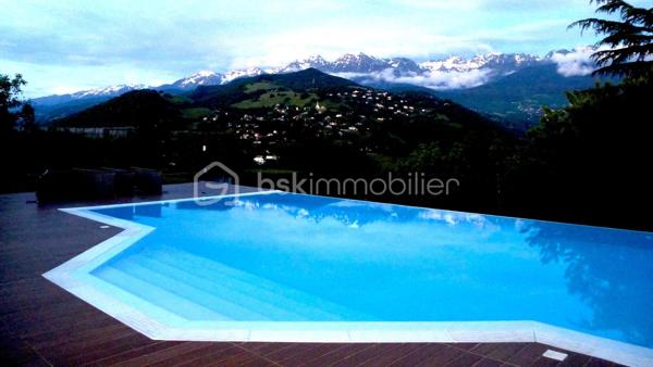 bonjour. Voici l'essentiel : 
à Gières au fort du Mûrier, une villa-belvédère 
258 m2 habitables 
158 m2 locaux annexes
416 m2 surface utile
6 chambres
4 salles de bain
Piscine à débordement
Terrain arboré et clos : 6737 m2 (chemin de ronde partiellement)

Bienvenue à celles et ceux qui préfère emprunter le « chemin de ronde » architectural :
A l'aube des années 80, aucune entrave à l'audace de ces jeunes architectes et commanditaires qui saisissent cette exceptionnelle opportunité de construire cette villa-belvédère au flanc du Fort du Mûrier à Gières qui verrouille la vallée du Grésivaudan.
La villa lui emprunte sa matérialité, son ancrage et sa compacité qu'elle exprime par les outils architecturaux développés dans le sillage de Le Corbusier.
Là où le Fort retourne comme un gant l'alignement des façades de ses casemates en une rue utilitaire  encastrée, la villa, sous le ruban de la haute poutre-acrotère du toit-terrasse, déploie, par redents irréguliers, l'alternance de ses parois pleines et vitrées toute hauteur.
Elle exalte la minéralité de sa périphérie de terrasses gravillonnées, d'une brique nuancée appareillée horizontalement en pans ourlés et d'un béton aux épidermes multiples tantôt lisse, tantôt brut de décoffrage, parfois avec méplats mais toujours d'expression verticale et favorise sa vocation résidentielle par les nombreux et généreux pans vitrés de son rez-de-jardin de 222 m2 intégralement de plain-pied. 
A cette projection dans le paysage proche et lointain répond une fluidité intérieure autorisée par la prédilection des échappées visuelles obliques et un certain dépouillement général.
Au final,  sans en imposer mais avec fermeté, sans mimétisme au fort, par son inaltérable minimalisme, la villa-belvédère semble surgie hier autant que depuis des temps immémoriaux.

Une chambre d'écho aux bruissements des éléments, du végétal et de la montagne.
Comme le fort organise sa vie défensive en fonction de sa rue en boomerang, la villa répartit son organisation de part et d'autre d'une rue intérieure dotée de la même orientation nord-ouest/sud-est salutaire eu égard au réchauffement climatique. 
Au nord, l’espace nuit de hauteur conventionnelle comprend 4 chambres, 3 salles d’eau nimbées d'une subtile lumière zénithale et 2 wc. Toutes les chambres s'exonèrent d'une vue frontale sur le fort au profit d'orientations est et ouest plus agréables. La suite parentale outre le privilège de ses 27 m2 et sa baie cadrant les trois pics de Freydone, dispose d'un accès doublé par le salon surélevé. Arrêtons-nous sur cette pièce singulière qui jouit d'un rapport visuel immédiat avec le miroir d'eau sur lequel flotte la chaine de Belledonne...salon tour-à-tour empli de la rumeur ludique de la piscine et assurément contemplative la nuit ou aux aurores, sa triple ouverture et son surplomb du hall l'apparentent au campement à la « croisée des chemins »  ce dont atteste la ligne fuyant et sinueuse des ganivelles de châtaigner du chemin de ronde.

Au sud de la rue intérieure, avec laquelle ils partagent une hauteur sous plafond avoisinant les 3 mètres, se déploient les lieux de rassemblement ou de concentration que sont le séjour, la cuisine, la salle-à-manger et le bureau.
Le séjour en contrebas de la rue intérieure s'organise autour du bloc massif intégrant le foyer-insert et son alter-ego le barbecue de la terrasse couverte des repas de mi-saison et d'été. Son plafond de panneaux de chêne massif confère à la pièce une couleur acoustique propice aux confidences.
On peut qualifier la cuisine d'agora familiale tant son organisation centripète permet de s'y tenir bien au-delà de sa fonction première de préparation des repas. Sa table tout en transparence dissout la frontière entre cuisine et terrasse tandis que ses ouvertures cardinales en diagonale permettent un contrôle visuel de toute la propriété : l'arrivée des visiteurs, le chevreuil du chemin de ronde, le gazouillis des oiseaux proches. Et que dire de son mobilier minimaliste et inaltérable aux couleurs hédoniques qui répond à sa mission depuis un demi-siècle avec la même justesse ?
Le niveau bas -d'égale surface- est purement fonctionnel. Au nord-ouest et de plain-pied avec l'accès à la propriété, le vaste garage est accessible par 3 portes sectionnelles et donne accès à un studio. Du fait de l'encastrement dans la déclivité du terrain et donc totalement enterrés, vous trouvez les espaces techniques tels que -principalement- la filtration de la piscine à débordement, la chaufferie et sa chaudière Weishaupt, la conserverie-cave à vins, la buanderie, l'atelier. Les vides sanitaires hauteur d'homme et partiellement soustrait à la roche réservent des opportunités d'aménagement. 

La villa s'implante au sud de l'équerre formé par le chemin de ronde et en lisière du hameau -solitaire mais solidaire- dont elle se protège par un écran végétal et une certaine déclivité.
Prolongement naturel du demi-kilomètre que constitue le tour de la propriété, un escalier extérieur donne accès au toit-terrasse qui offre une vue panoramique à 360 degrés sur le Vercors, Grenoble, la dent de Crolles, le Grésivaudan, le Mont-blanc par beau temps et la chaine de Belledonne. 
La boucle du Fort du Mûrier passe au pied de la propriété et offre au cours de ses deux heures de marche une succession de panoramas, selon les randonneurs, « à coupe le souffle ».
Aux portes de Grenoble, Gières, bien desservie par la rocade nord-est et à la croisée de plusieurs domaines skiables rejoints en moins d'une demie-heure, elle est dotée de tous les commerces et services. Mais une telle adresse se mérite et il faut - depuis le bourg - gravir un dénivelé de plus de 200 mètres pour atteindre le Fort et la villa.

Alain CHIAUDANO & Régis HERBIN, l’héroïque binôme architectural 
Durant les deux années précédant la construction, les commanditaires ont affronté de nombreuses résistances à leur projet mais cela n'a entamé en rien ni leur envie de vivre en ce lieu solitaire et grandiose ni la conviction d'Alain Chiaudano d'un plateau de vie -« bunker » en lévitation- affranchi de toute enveloppe conventionnelle. Malheureusement, quelques mois avant l'ouverture du chantier, l'architecte est emporté par la maladie et c'est son ami et associé régis Herbin qui fera preuve d'abnégation et de respect mémoriel. Porté par l'encouragement sans faille des commanditaires, le jeune architecte livrera en 15 mois une œuvre unique où tout est calepiné, apparent, doté d'une science des rythmes, des épidermes et de l'espace qui n'est pas sans rappeler l'Atelier de Montrouge de la première heure ou le binôme Le Corbusier-Wogenscky. 
Les améliorations que les résidents lui ont prodigué au fil des ans, la belle patine des ouvrages et au final, l'absence de toute modification majeure témoignent de la pertinence des choix initiaux et attestent de son état impeccable.