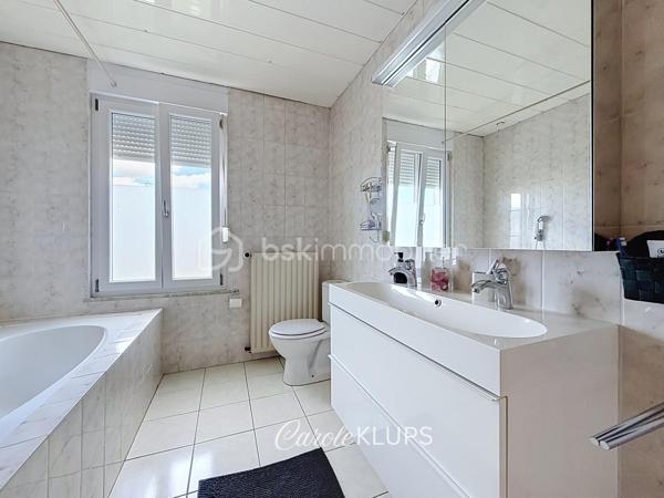 Maison familiale avec véranda, terrasse couverte, jardin et piscine à Village-Neuf

Ici, la surface habitable ne raconte pas tout. 112 m² habitables auxquels s’ajoutent une véranda d’environ 10 m², une terrasse couverte tournée vers le jardin, un sous-sol et un double garage. 

Le charme d’une maison des années 30 dans laquelle on peut se créer une véritable vie dedans-dehors, à deux pas de la frontière bâloise. Et pour les frontaliers, Bâle est accessible à vélo via la piste cyclable longeant le canal de Huningue.

Depuis la rue, on ne soupçonne pas le cadre de vie que cette maison réserve à l’arrière.

Résolument tournée vers l’extérieur, elle s’ouvre sur un jardin intimiste où chaque espace trouve naturellement sa place. Une belle terrasse couverte pour les repas entre amis ou en famille. À moins que vous ne préfériez vous installer au bord de la piscine chauffée … Autour, l’espace paysager invite au repos et à la convivialité.

Une sensation de vacances chez soi, avec un autre avantage appréciable : un jardin aux dimensions raisonnables, pensé pour en profiter sans devenir esclave de son entretien. Un puits présent sur la propriété permet par ailleurs d’assurer l’arrosage du jardin.

À l’intérieur aussi, chaque espace a sa fonction et la circulation est fluide.

Au rez-de-chaussée, le séjour de 32,6 m² invite à se prélasser près du poêle à bois. La cuisine équipée de 12 m² offre un espace confortable pour cuisiner, tandis que la véranda d’environ 10 m² devient naturellement une pièce supplémentaire pour partager les repas, avec le jardin en toile de fond.

Le + de ce rez-de-chaussée ? Une salle d’eau avec WC.

En montant à l’étage, même le palier a été optimisé avec un petit espace buanderie astucieusement dissimulé.

Puis vient l’une des belles surprises de la maison : une vaste mezzanine de plus de 23 m².

Aujourd’hui ouverte, elle sera votre terrain de jeu. Espace télétravail, bibliothèque, salle de jeux pour les enfants, salon télé… à chacun d’imaginer sa façon de l’habiter. Elle pourra également être cloisonnée pour créer une troisième chambre, qui viendra alors compléter les deux chambres existantes de 12,6 et 11,2 m².

La salle de bains de 7,6 m², avec baignoire, double vasque et WC, est quant à elle dimensionnée pour ces matins en famille où l’on apprécie de ne pas se marcher dessus.

Et ce n’est pas tout.

Au sous-sol, on retrouve une partie chaufferie, un atelier ainsi qu’un espace déjà aménagé en salle de sport, à conserver tel quel ou à réinventer selon vos besoins.

À l’extérieur, le double garage, la piscine chauffée et son local technique indépendant complètent les prestations de la maison.

Le tout prend place sur une parcelle de 480 m².

Enfin, sa situation en zone frontalière, avec des accès faciles vers la Suisse comme vers l’Allemagne, est particulièrement adaptée à la vie quotidienne des actifs du secteur.

Une maison avec jardin et piscine, une vraie vie dedans-dehors et Bâle à portée de vélo : profiter pleinement de chez soi sans renoncer à la proximité de la frontière.