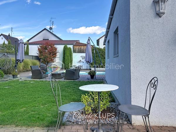 Maison familiale avec véranda, terrasse couverte, jardin et piscine à Village-Neuf

Ici, la surface habitable ne raconte pas tout. 112 m² habitables auxquels s’ajoutent une véranda d’environ 10 m², une terrasse couverte tournée vers le jardin, un sous-sol et un double garage. 

Le charme d’une maison des années 30 dans laquelle on peut se créer une véritable vie dedans-dehors, à deux pas de la frontière bâloise. Et pour les frontaliers, Bâle est accessible à vélo via la piste cyclable longeant le canal de Huningue.

Depuis la rue, on ne soupçonne pas le cadre de vie que cette maison réserve à l’arrière.

Résolument tournée vers l’extérieur, elle s’ouvre sur un jardin intimiste où chaque espace trouve naturellement sa place. Une belle terrasse couverte pour les repas entre amis ou en famille. À moins que vous ne préfériez vous installer au bord de la piscine chauffée … Autour, l’espace paysager invite au repos et à la convivialité.

Une sensation de vacances chez soi, avec un autre avantage appréciable : un jardin aux dimensions raisonnables, pensé pour en profiter sans devenir esclave de son entretien. Un puits présent sur la propriété permet par ailleurs d’assurer l’arrosage du jardin.

À l’intérieur aussi, chaque espace a sa fonction et la circulation est fluide.

Au rez-de-chaussée, le séjour de 32,6 m² invite à se prélasser près du poêle à bois. La cuisine équipée de 12 m² offre un espace confortable pour cuisiner, tandis que la véranda d’environ 10 m² devient naturellement une pièce supplémentaire pour partager les repas, avec le jardin en toile de fond.

Le + de ce rez-de-chaussée ? Une salle d’eau avec WC.

En montant à l’étage, même le palier a été optimisé avec un petit espace buanderie astucieusement dissimulé.

Puis vient l’une des belles surprises de la maison : une vaste mezzanine de plus de 23 m².

Aujourd’hui ouverte, elle sera votre terrain de jeu. Espace télétravail, bibliothèque, salle de jeux pour les enfants, salon télé… à chacun d’imaginer sa façon de l’habiter. Elle pourra également être cloisonnée pour créer une troisième chambre, qui viendra alors compléter les deux chambres existantes de 12,6 et 11,2 m².

La salle de bains de 7,6 m², avec baignoire, double vasque et WC, est quant à elle dimensionnée pour ces matins en famille où l’on apprécie de ne pas se marcher dessus.

Et ce n’est pas tout.

Au sous-sol, on retrouve une partie chaufferie, un atelier ainsi qu’un espace déjà aménagé en salle de sport, à conserver tel quel ou à réinventer selon vos besoins.

À l’extérieur, le double garage, la piscine chauffée et son local technique indépendant complètent les prestations de la maison.

Le tout prend place sur une parcelle de 480 m².

Enfin, sa situation en zone frontalière, avec des accès faciles vers la Suisse comme vers l’Allemagne, est particulièrement adaptée à la vie quotidienne des actifs du secteur.

Une maison avec jardin et piscine, une vraie vie dedans-dehors et Bâle à portée de vélo : profiter pleinement de chez soi sans renoncer à la proximité de la frontière.